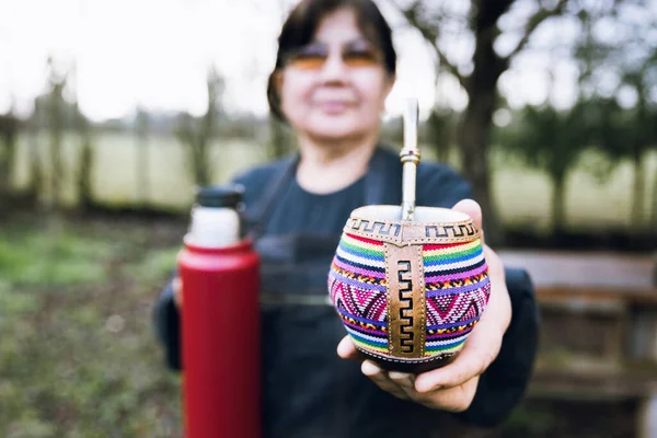 Latin woman serving mate with a red thermo, in a colorful ethnic mate. Latin beverage. Ethnical concept. High quality photo.