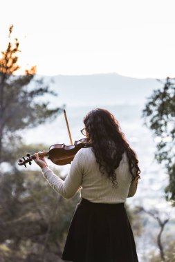 Brunette woman seen from back playing violin outside in the mountain. Vertical. High quality photo