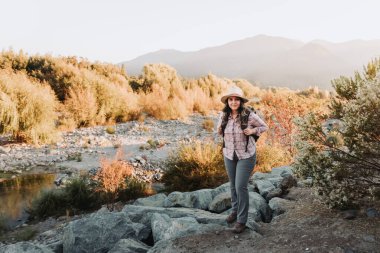 Güzel bir manzarada yürüyüş yapan şapkalı ve sırt çantalı genç Latin bir kadın. Yaz havası. Yüksek kalite fotoğraf. Boşluğu kopyala.