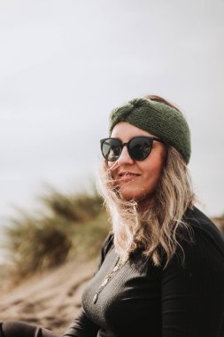 Smiling blonde woman dressed in black, wearing a green headband, sunglasses and sitting on the beach. High quality photo