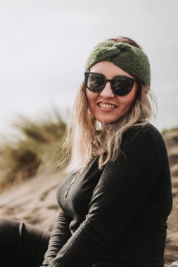 Smiling blonde woman dressed in black, wearing a green headband, sunglasses and sitting on the beach. High quality photo