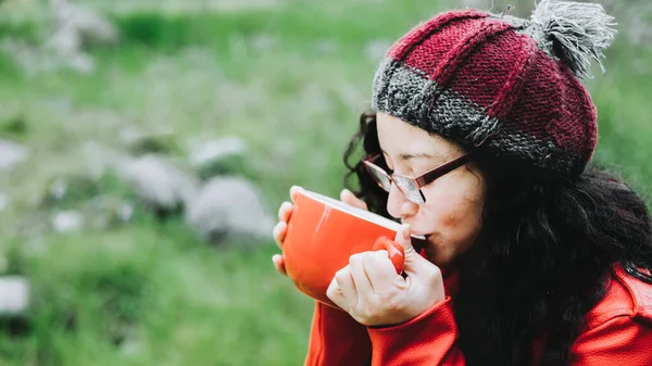 Curly brunette woman, wearing a red leather jacket, and holding a red mug with hot coffee in nature. High quality photo