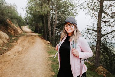 Tired young latin plus size woman with a hat and a backpack on, hiking in a beautiful landscape. High quality photo. Copy space.