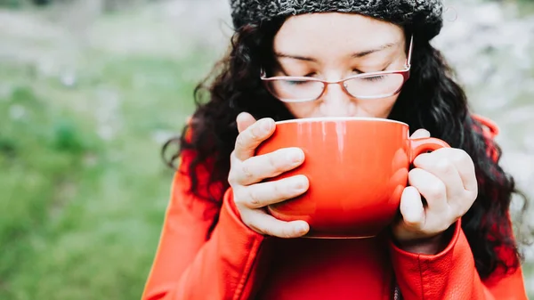 Curly brunette woman, wearing a red leather jacket, and holding a red mug with hot coffee in nature. High quality photo