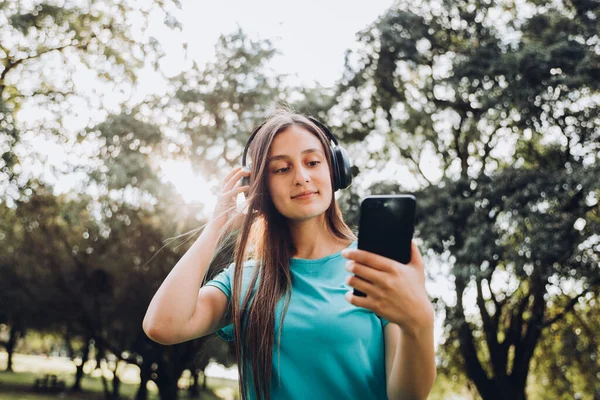 Young girl wearing turquoise t shirt using black headphones and setting a music playlist on her smartphone in the park. High quality photo