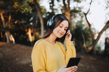 Sarı kazak giyen genç kız, parkta akıllı telefonuyla müzik çalmak için kulaklık kullanıyor. Yüksek kalite fotoğraf