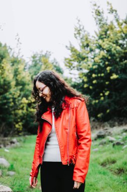 Curly brunette woman, wearing a red leather jacket, and holding a red mug with hot coffee in nature. High quality photo