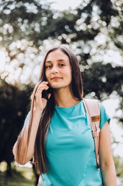 Turkuaz tişört giyen genç kız parkta akıllı telefonuyla bir telefon görüşmesi yapıyor. Güneş ışığı. Yüksek kalite fotoğraf