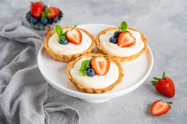 Taze böğürtlenli lezzetli mini tartlar ve gri beton arka planda beyaz bir tabakta krema. Boşluğu kopyala.