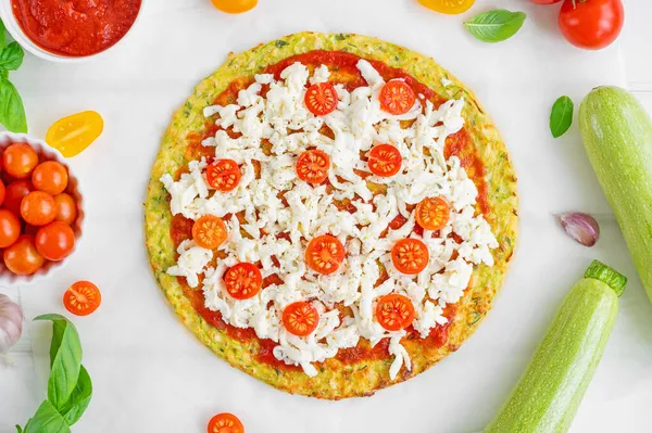 Preparation zucchini crust pizza with tomato sauce, mozzarella cheese, fresh tomatoes and basil on a white wooden background. Zucchini pizza Margarita. Healthy dish
