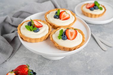 Taze böğürtlenli lezzetli mini tartlar ve gri beton arka planda beyaz bir tabakta krema. Boşluğu kopyala.