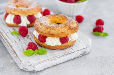 Choux hamur işleri. Krem peynirli Choux halkaları veya süzme peynir ve taze ahududu tozlu tahta bir tahta üzerinde gri beton arka planda toz şeker