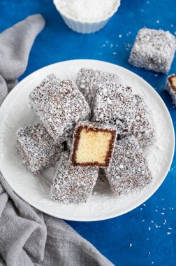 Geleneksel Avustralya Lamington pastası çikolata jöleli ve beyaz bir tabakta hindistan cevizi gevrekleri ve bir fincan çay eşliğinde mavi beton arka planda. Seçici odak