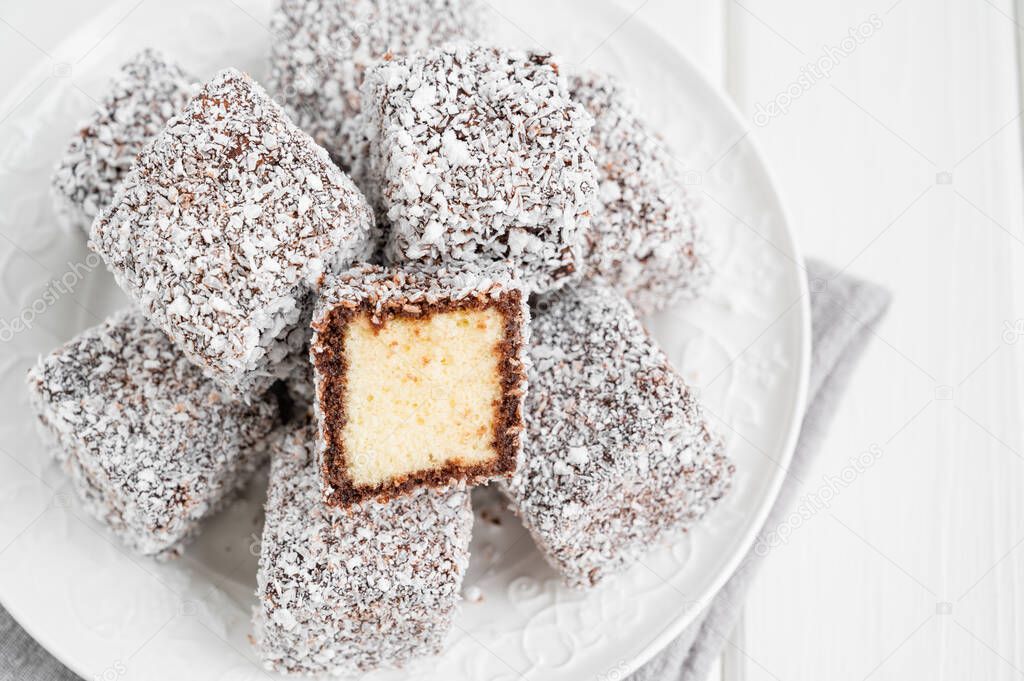 Traditional Australian Lamington cake in chocolate icing and coconut chips on a white plate on a white wooden background with a cup of tea. Selective focus
