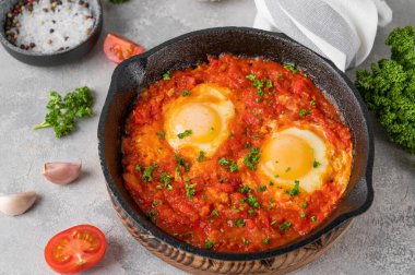 Shakshuka kızartma tavasında gri beton arka planda malzemelerle birlikte. Baharatlı ve taze maydanozlu domates soslu yumurta. Boşluğu kopyala