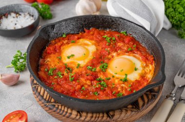 Shakshuka kızartma tavasında gri beton arka planda malzemelerle birlikte. Baharatlı ve taze maydanozlu domates soslu yumurta. Boşluğu kopyala