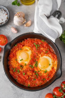 Shakshuka kızartma tavasında gri beton arka planda malzemelerle birlikte. Baharatlı ve taze maydanozlu domates soslu yumurta. Boşluğu kopyala