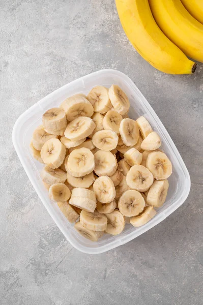 Slices of frozen banana in a container on a gray concrete background. Ingredient for smoothies, ice cream, desserts. Healthy food. Top view