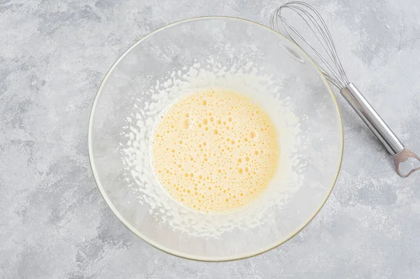 Raw beaten eggs with sugar in a glass bowl on a gray concrete background. Preparation of cream, scrambled eggs, French toasts, desserts. Top view