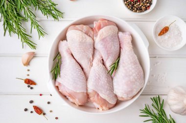 Raw uncooked chicken legs in a white dish on a white wooden background. Meat with ingredients for cooking. Top view