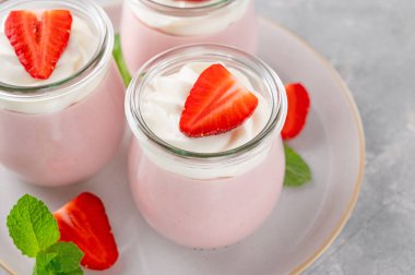 Strawberry mousse in a glass jar on a gray concrete background with whipped cream, fresh berries and mint leaves. Delicious summer dessert