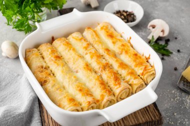 Pancakes or thin crepes stuffed with chicken and mushrooms baked with cheese in a baking dish on a gray concrete background.