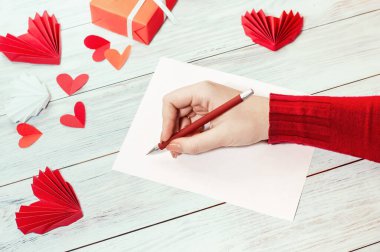 a girl writes a congratulatory love letter on a bright table with red symbols of love and a gift