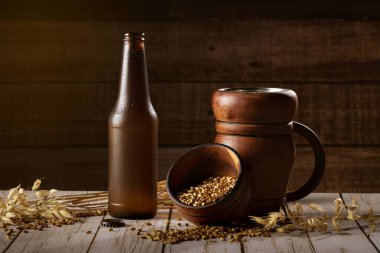 Bottle of cold beer placed on a rustic wooden table with a bowl of barley grains. Dark background