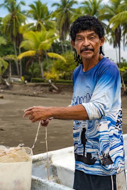 Kameraya bakan adam, arka planda palmiye ağaçları olan teknesinde balık tutmak için ağını hazırlıyor..