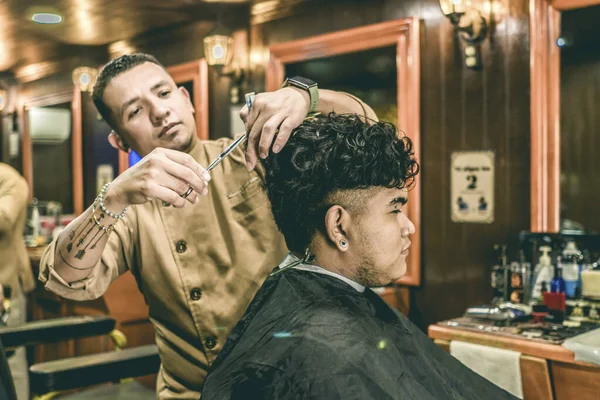 Serious tattooed barber cutting hair of young man with piercings sitting in a chair in a barber shop.