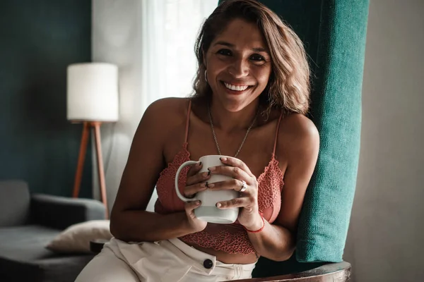 Happy young Latina woman smiles looking at the camera, holding a cup and enjoying a new day. Healthy lifestyle concept.