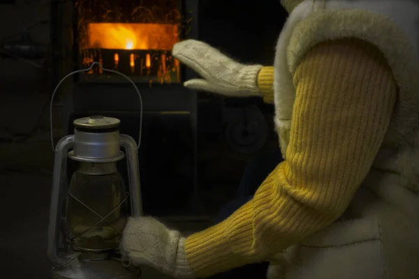 Starsza kobieta w ciepłym swetrze i kamizelce ogrzewa swoją rękawicę przy kominku w piekarniku, a drugą trzyma starą lampę naftową. Zimna zima, ogrzewanie zimą, brak prądu — Zdjęcie stockowe