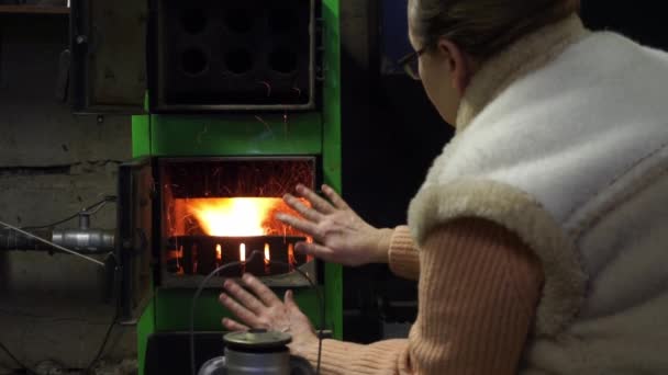 Une femme âgée vêtue d'un pull et d'un gilet chauds étend ses paumes au feu dans un four à charbon, se réchauffe par le feu. Hiver froid, chauffage au charbon en hiver, pas d'électricité — Video