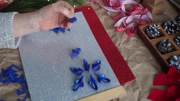 女性の手は、テーブルのカラフルなファミランシート、赤と白のリボン、コーンの上に、青い輝きのファミランから雪の結晶要素をカットしました。クリスマスの到来のカレンダーの準備。DIY — ストック動画
