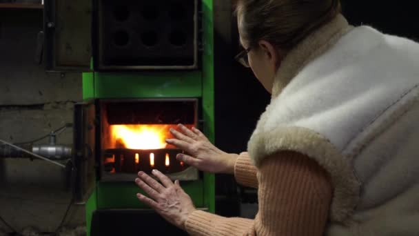 Une femme âgée vêtue d'un pull et d'un gilet chauds étend ses paumes au feu dans un four à charbon, se réchauffe par le feu. Hiver froid, chauffage au charbon en hiver, pas d'électricité — Video
