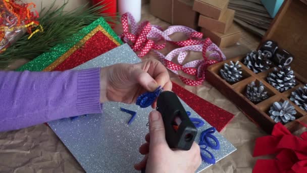 De vrouwenhanden knippen het sneeuwvlokkenelement uit blauwe glitterschuim, op de tafel kleurrijke schuimiran lakens, rood en wit lint, kegels. Ik bereid me voor op Kerstmis. DIY — Stockvideo