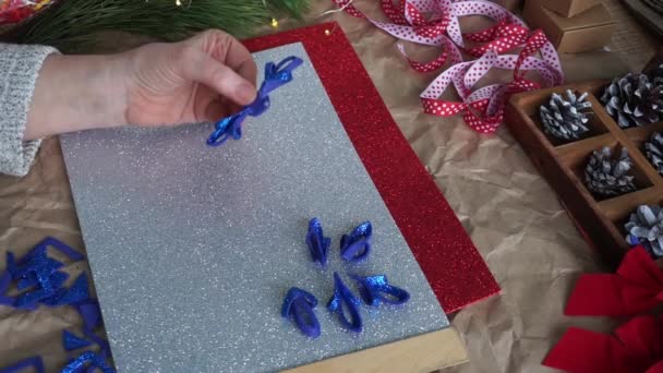 Le mani delle donne ritagliano l'elemento fiocco di neve da schiuma glitter blu, sul tavolo fogli di schiuma colorati, nastro rosso e bianco, coni. Prepararsi per il calendario dell'avvento di Natale. Fai da te — Video Stock