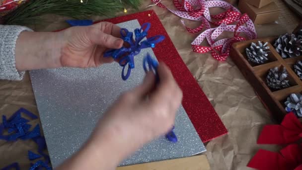 女性の手は、テーブルのカラフルなファミランシート、赤と白のリボン、コーンの上に、青い輝きのファミランから雪の結晶要素をカットしました。クリスマスの到来のカレンダーの準備。DIY — ストック動画