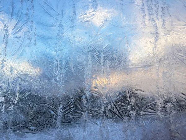 Krásným mrazivým vzorem na okenním skle prosvítí modrá obloha, mraky a domy. Přírodní textura — Stock fotografie