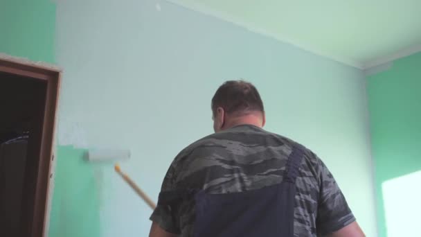 Starší muž v modré kombinéze maluje zelenou stěnu místnosti válečkem světle šedé barvy. Oprava konceptu, malování stěn vlastníma rukama — Stock video