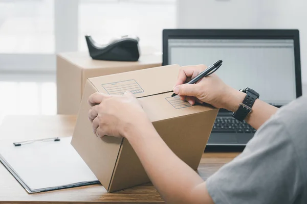 Close up hand a man use pen write on parcel box about the parcel delivery address and use laptop to register in the online system