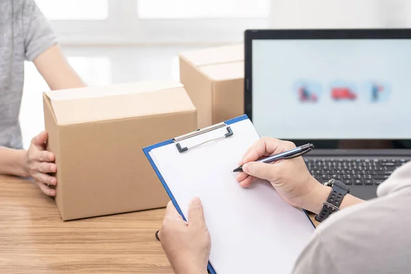 Post man use a pen write on file paper document and check online system to send parcels and women hold paper box laptop on table : delivery concept work