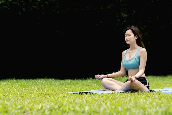 An asian woman in blue sport bra and black short sitting and doing meditation on blue yoga mat on green grass floor in garden with black and green blurry bokeh background with peaceful emotion.