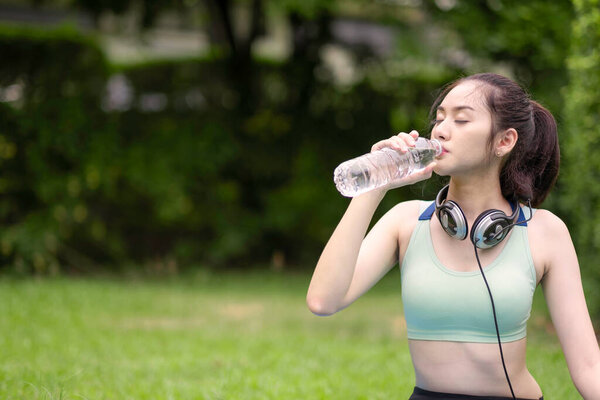 An asian gilr in green sport bra sitting and drinking water with closing eye after workout with green bokeh background at garden.
