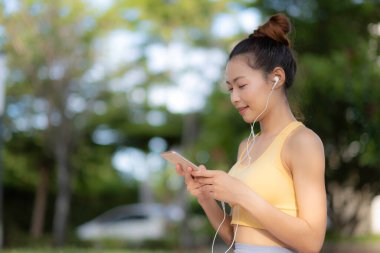Asyalı güzel bir kadın elinde gülümsemeli akıllı bir telefon, müzik dinleyen bir kadın ya da parkta kulaklıkla mutluluk sesi dinleyen bir kadın. Eğlence ve iyileşen zihin konsepti.