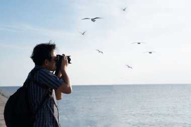 Mavi beyaz tişörtlü bir adam siyah çanta taşıyor ve okyanusta martı fotoğrafları çekiyor. Seyahat ediyorum ve fotoğraflarla hafızamı koruyorum.