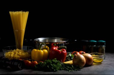 Spagetti içeriğinde kırmızı ve sarı dolma biber, soğan, domates, fesleğen ve siyah arka planda maydanoz vardı..