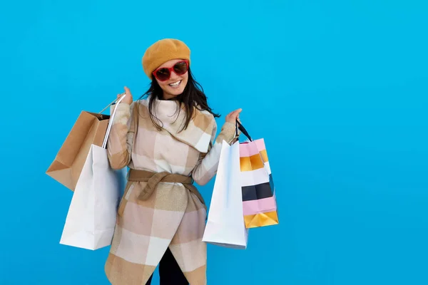トレンディーなベージュのコートとベレー帽を保持ショッピングバッグブルースタジオの背景にサングラスのお店でかわいい魅力的な幸せな笑顔スタイリッシュな女性独立したカラフルな販売興奮ファッショントレンド屋外 ストック画像
