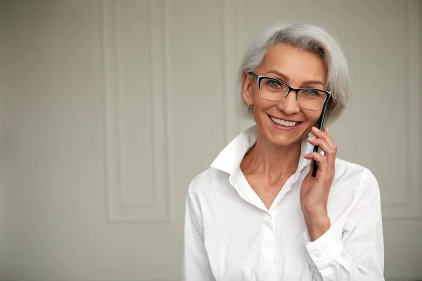 Cheerful mature businesswoman talking on smartphone white wall — Stock Photo, Image