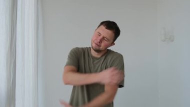 Pozitif adam beyaz duvarın yanında kameraya poz verirken kendini kucaklıyor.